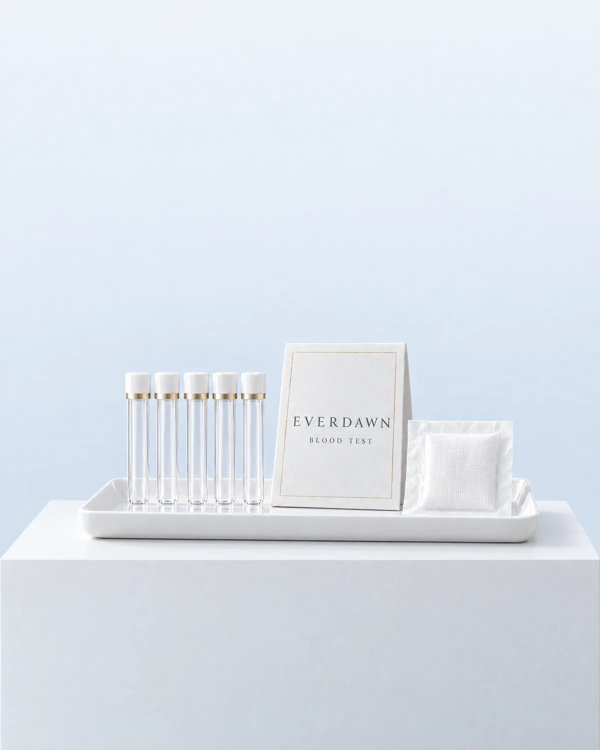 Empty blood collection tubes on a white tray beside an Everdawn card, on a white plinth in a pale blue studio.